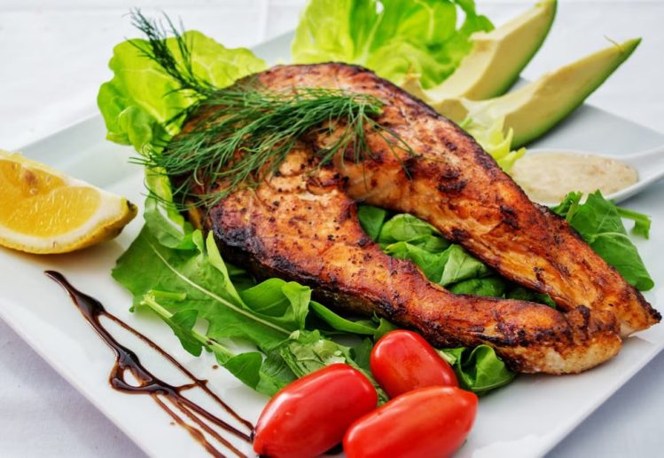 Delicious grilled salmon steak served with avocado, lettuce, cherry tomatoes, and lemon slice on a white plate.