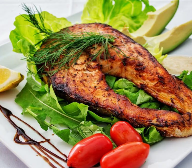 Delicious grilled salmon steak served with avocado, lettuce, cherry tomatoes, and lemon slice on a white plate.